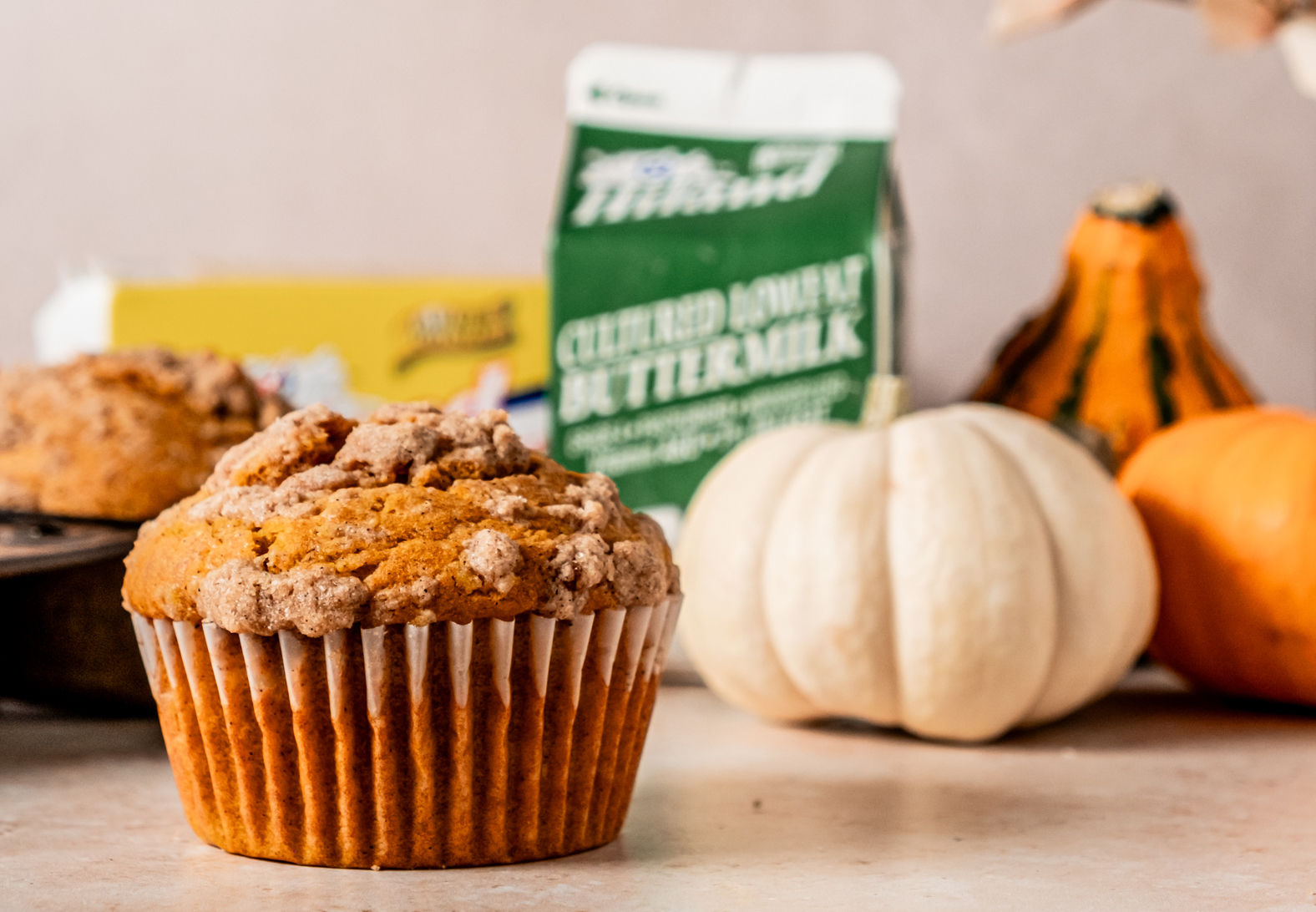 Another pumpkin streusel muffin with crumb topping, accompanied by Hiland buttermilk and butter in the background, alongside decorative pumpkins.