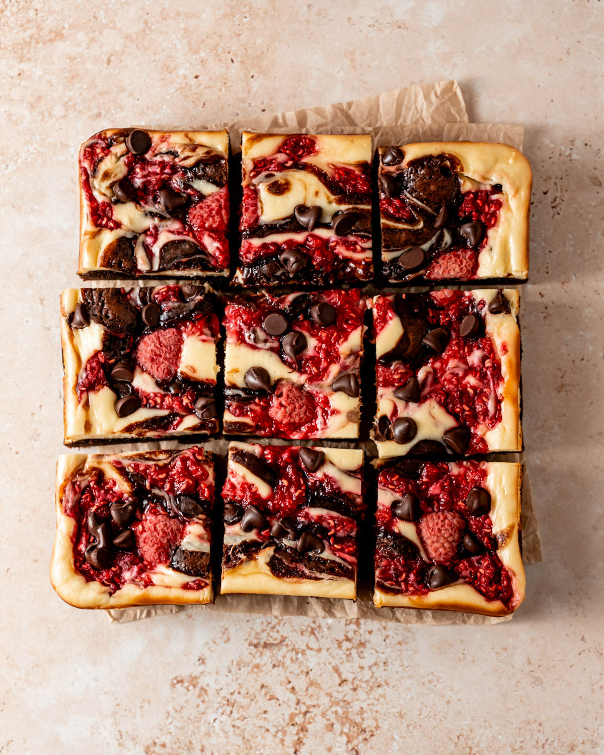 Sliced raspberry cheesecake brownies on parchment paper, showing vibrant layers of chocolate, cheesecake, and raspberries topped with chocolate chips.