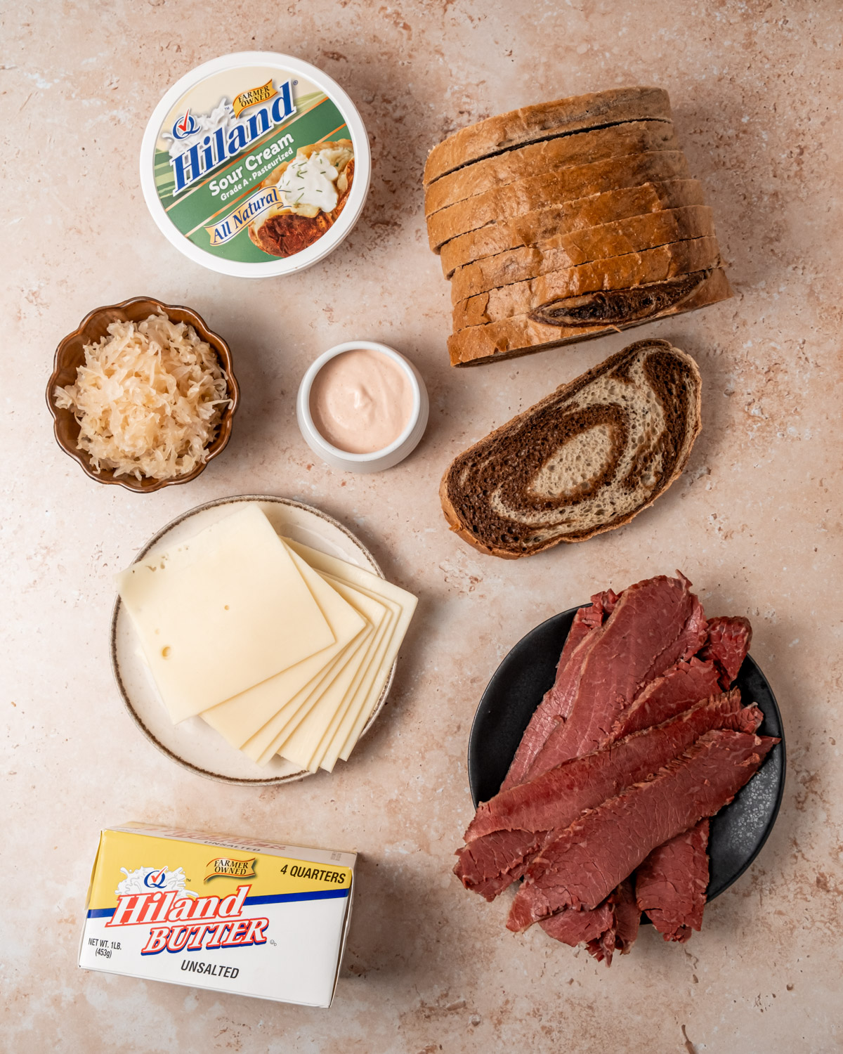 A top-down view of Reuben sandwich ingredients, including rye bread, Swiss cheese, corned beef, sauerkraut, and homemade Russian dressing. A container of Hiland Sour Cream and a package of Hiland Butter are also included in the display.