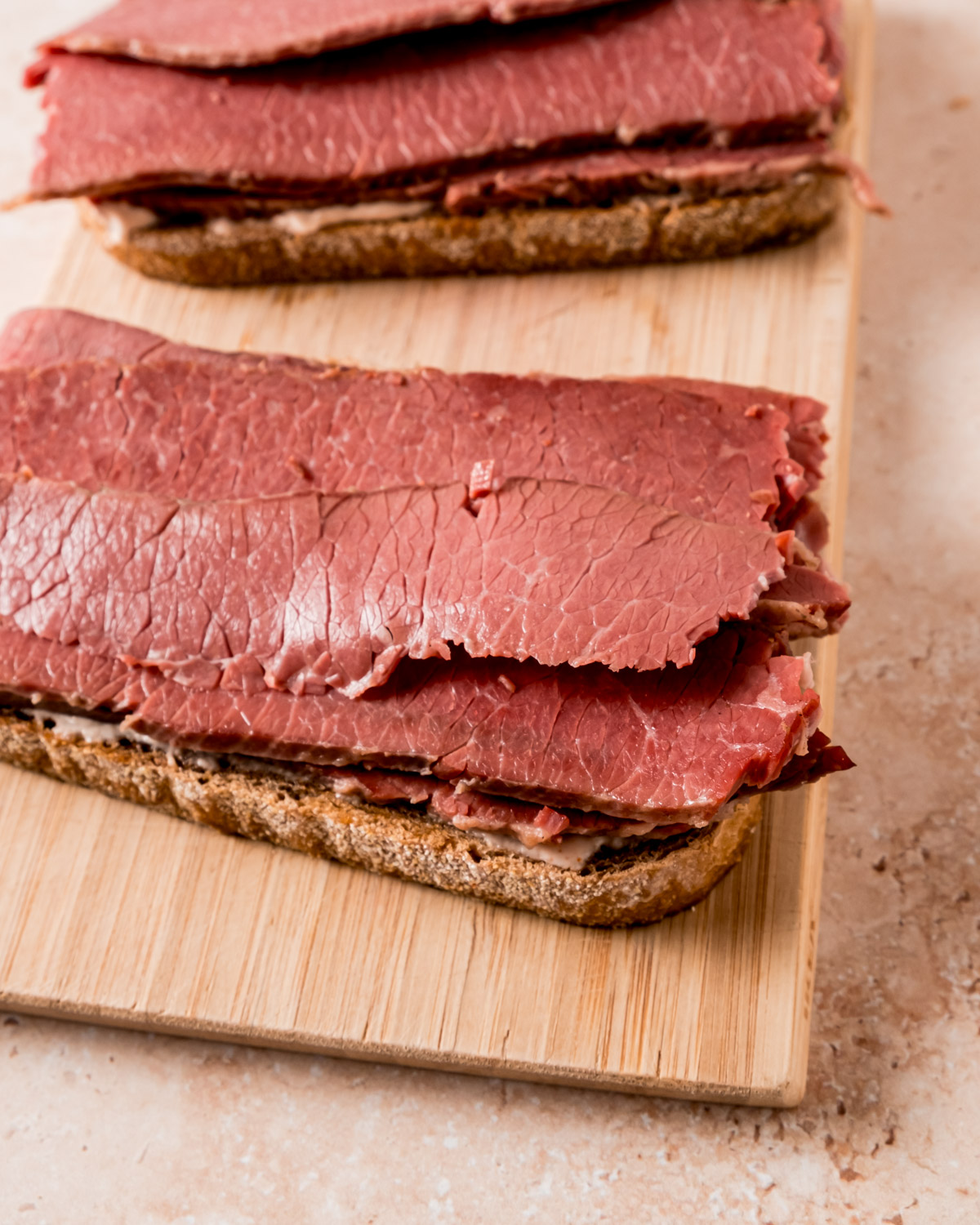 Open-faced sandwiches in progress on a wooden cutting board, showing slices of rye bread topped with layers of corned beef.