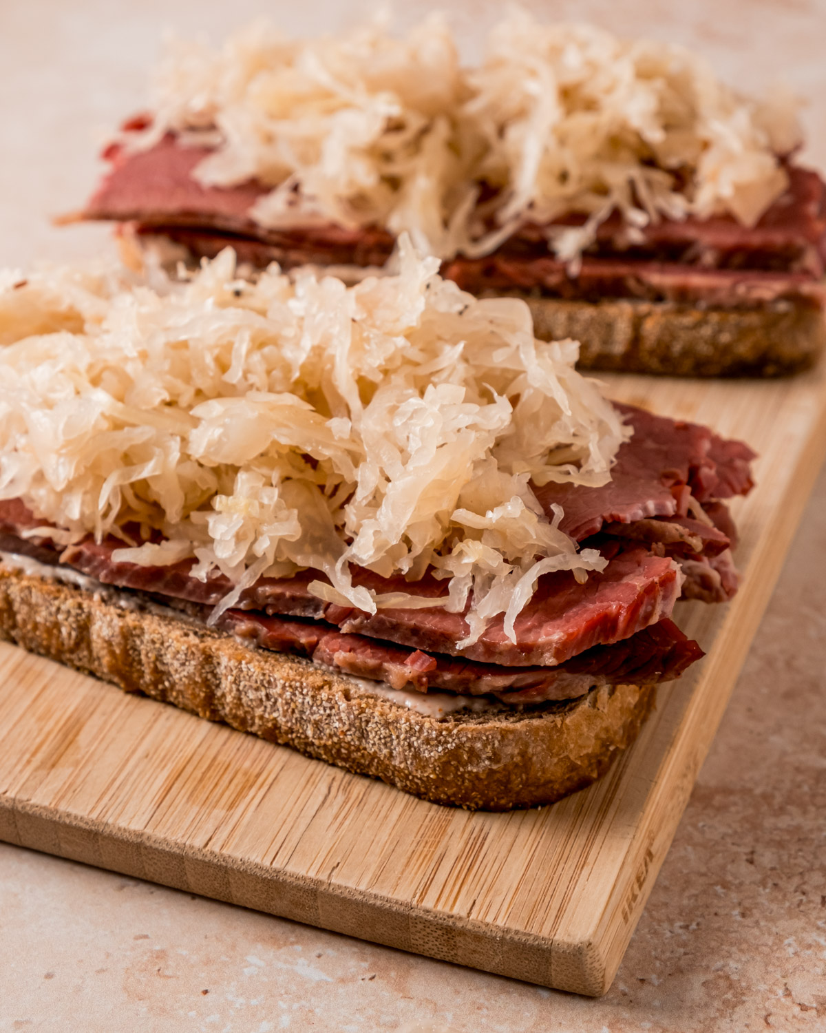 Open-faced Ruben sandwiches in progress on a wooden cutting board, topped with generous portions of sauerkraut, ready to be completed and grilled.