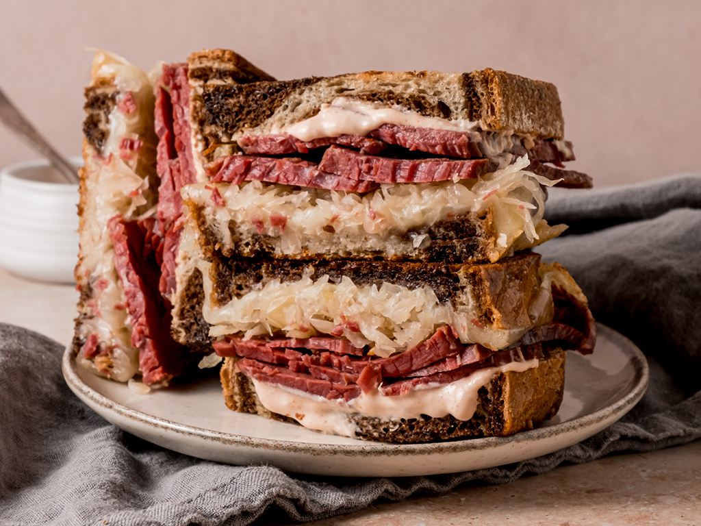 A close-up of a stacked Reuben sandwich revealing the rich layers of corned beef, sauerkraut, melted cheese, and dressing between toasted rye bread.