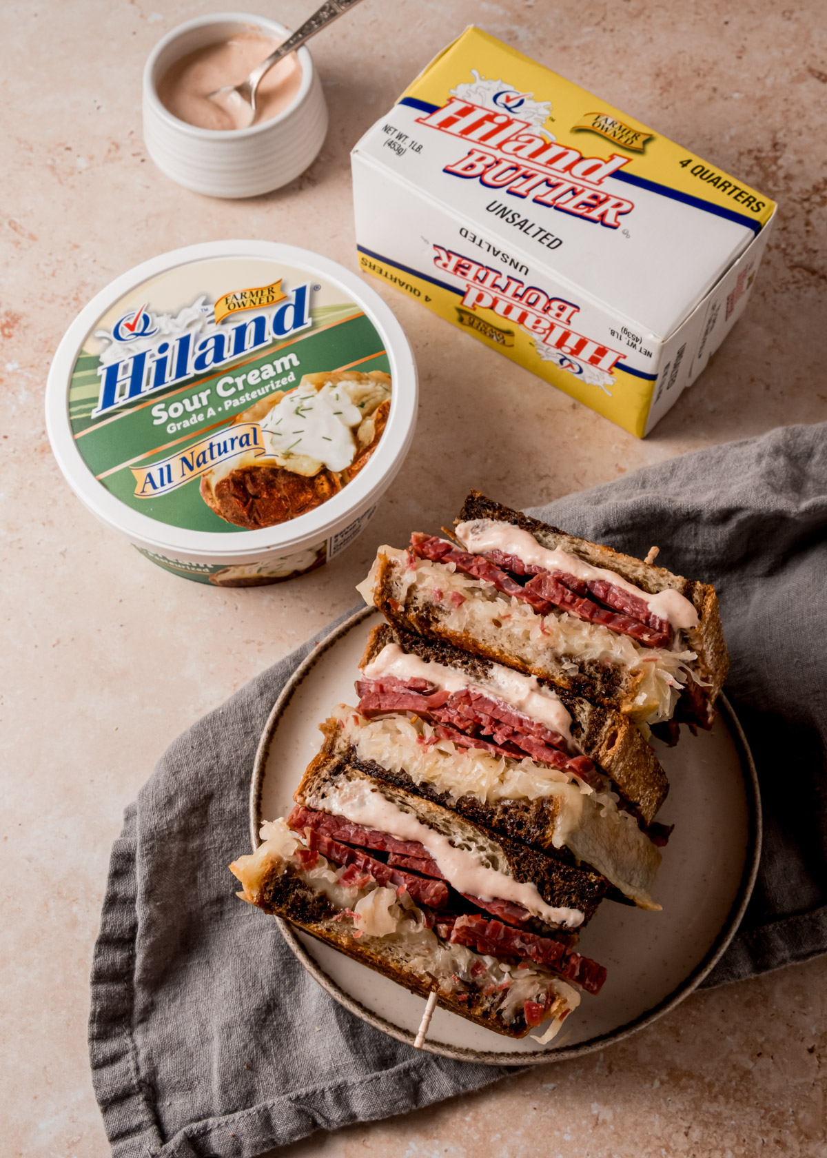 Vertically composed overhead view of a Reuben sandwich cut into halves on a plate that is on top of a gray cloth napkin, next to containers of Hiland Sour Cream and Hiland Butter, and a ramikin containing homemade Russian dressing.