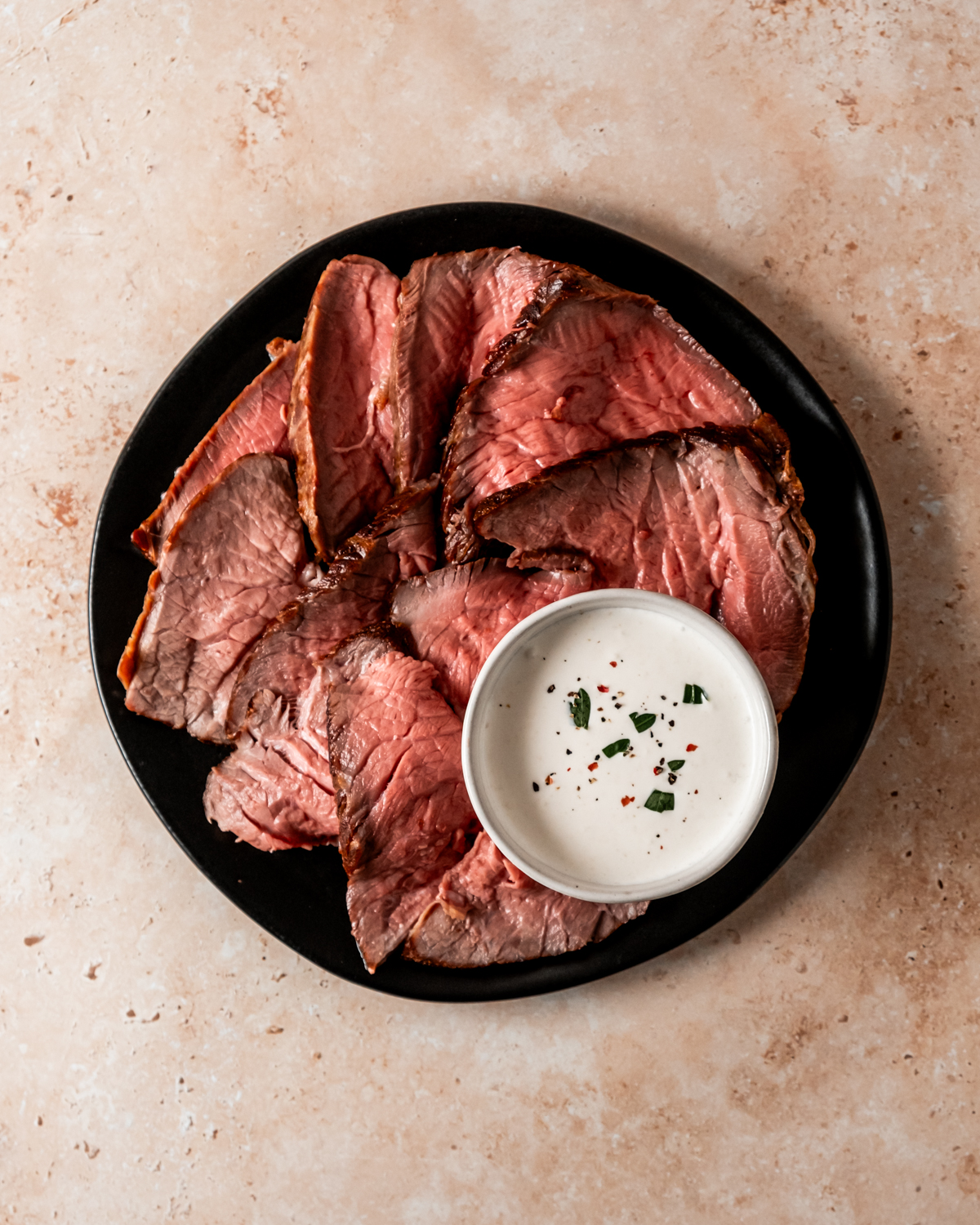 Slices of roast beef served on a black plate with a bowl of creamy sauce garnished with herbs.