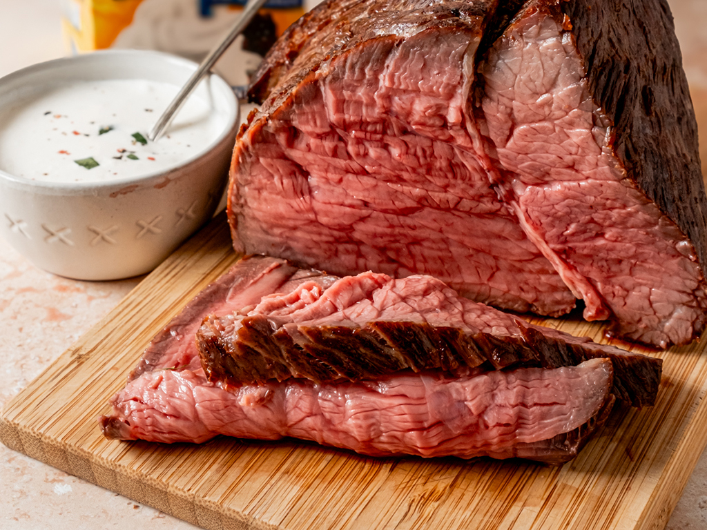 Close-up of a roast beef cut on a wooden board with slices in the foreground, next to a bowl of creamy sauce.