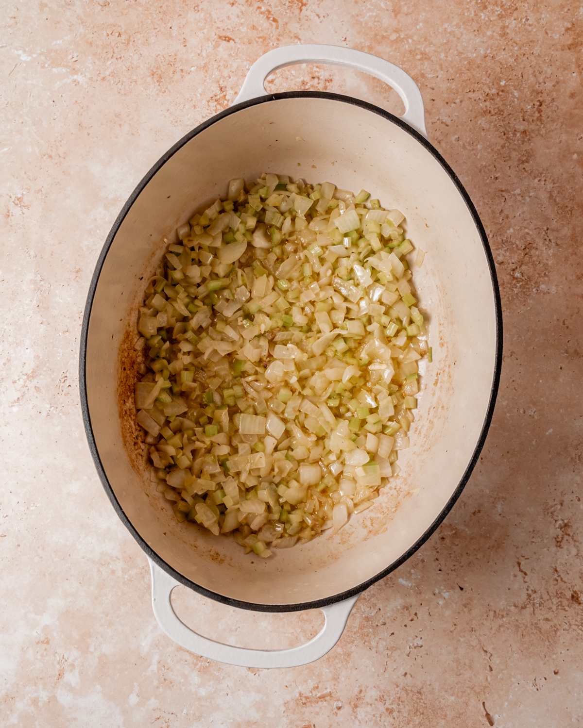 Diced onion and celery sautéing in the same white pot, softened and lightly golden, ready to be combined with other stuffing ingredients.