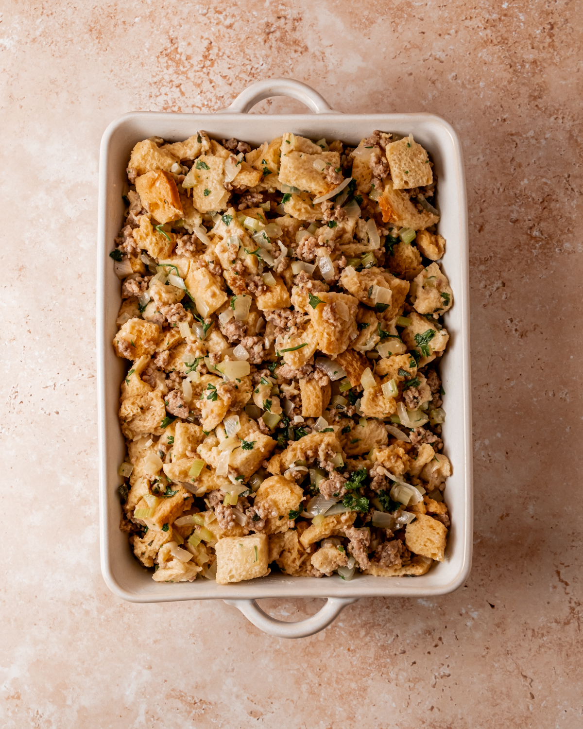 Sausage and sage stuffing mixture transferred into a white casserole dish, prepared for baking with a rustic texture and chunks of bread and vegetables.