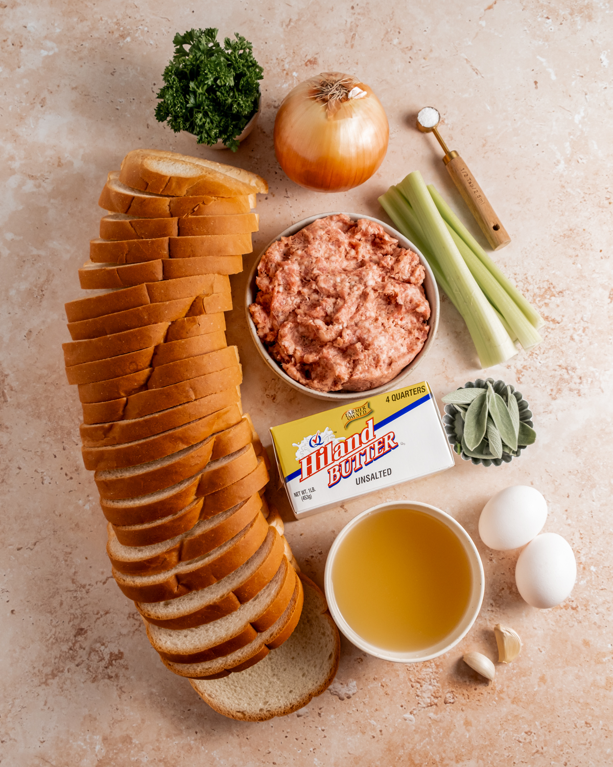 Ingredients for sausage and sage stuffing arranged on a countertop, including sliced bread, a bowl of ground sausage, Hiland butter, chopped parsley, whole onion, celery stalks, eggs, and a bowl of broth.