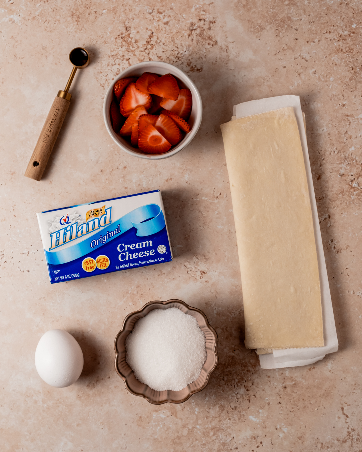 Ingredients for strawberry Danishes laid out on a light stone surface, including puff pastry, a bowl of sliced strawberries, an egg, sugar, and a package of Hiland Cream Cheese.