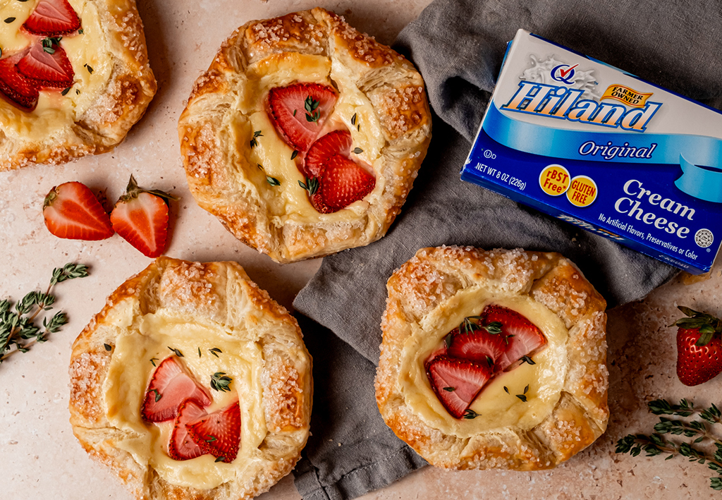 Several strawberry Danish pastries on a light surface with fresh strawberries, thyme sprigs, and a package of Hiland Cream Cheese, arranged on and around a gray cloth napkin.