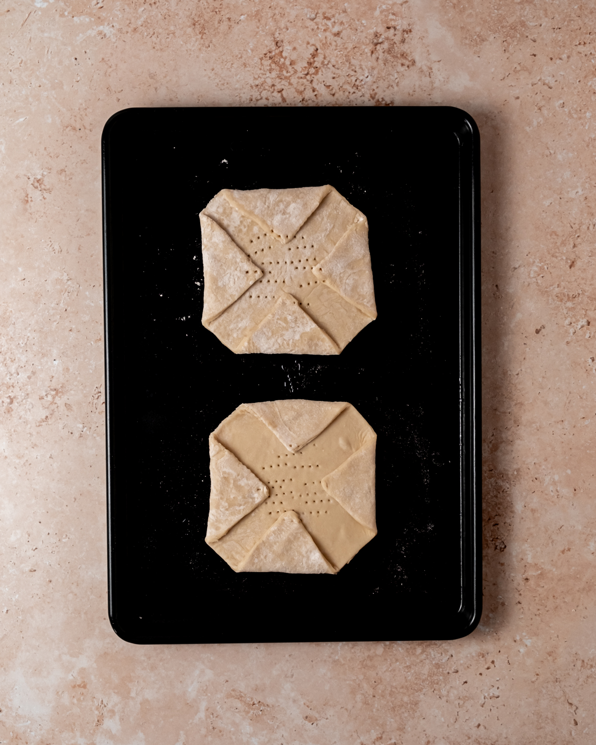 Two unbaked puff pastry squares folded into a pinwheel shape and docked with a fork, arranged on a dark baking tray, ready to be filled and baked.