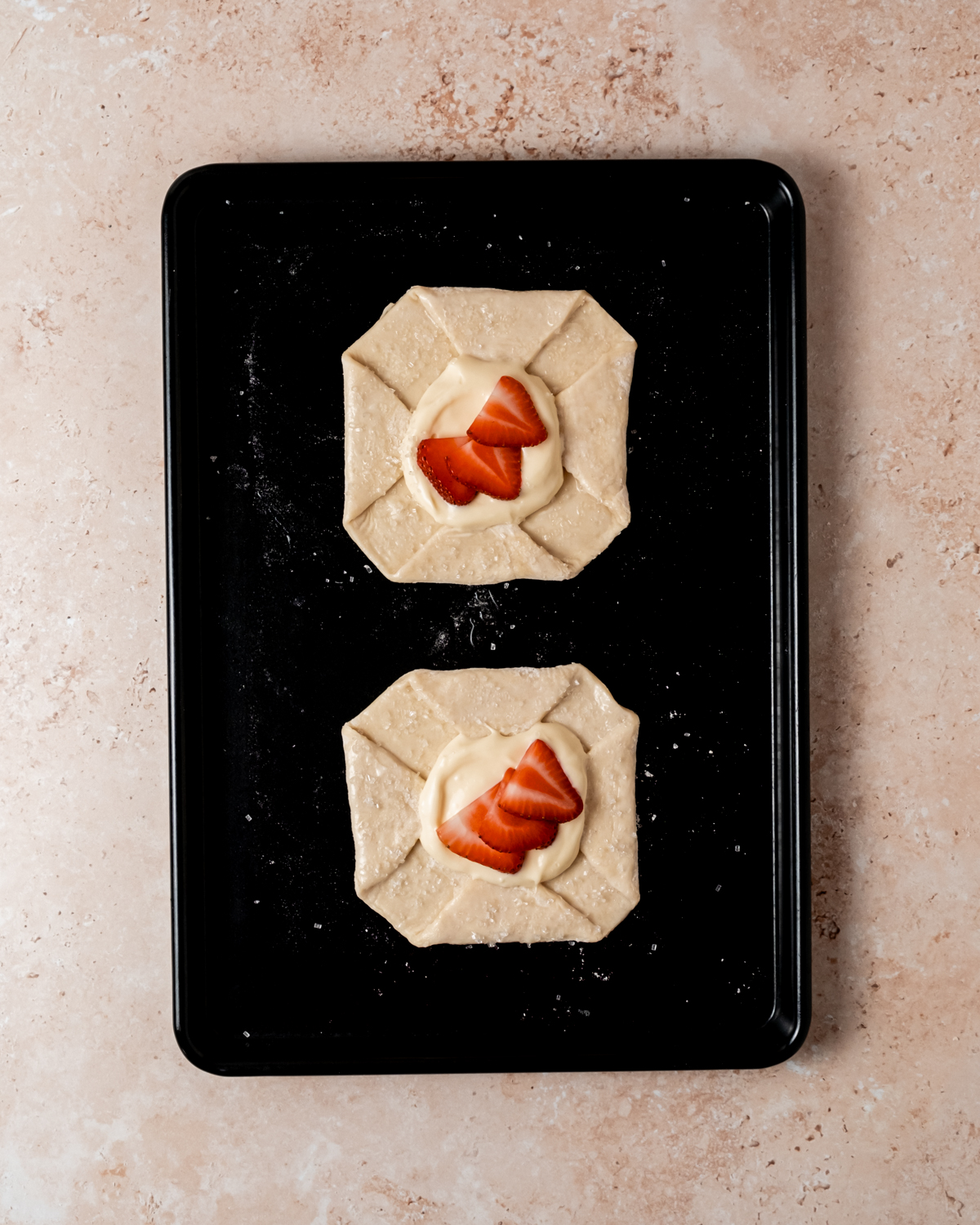 A top-down view of two unbaked strawberry danishes on a dark baking sheet, filled with cream cheese and topped with strawberry slices, on a speckled countertop.