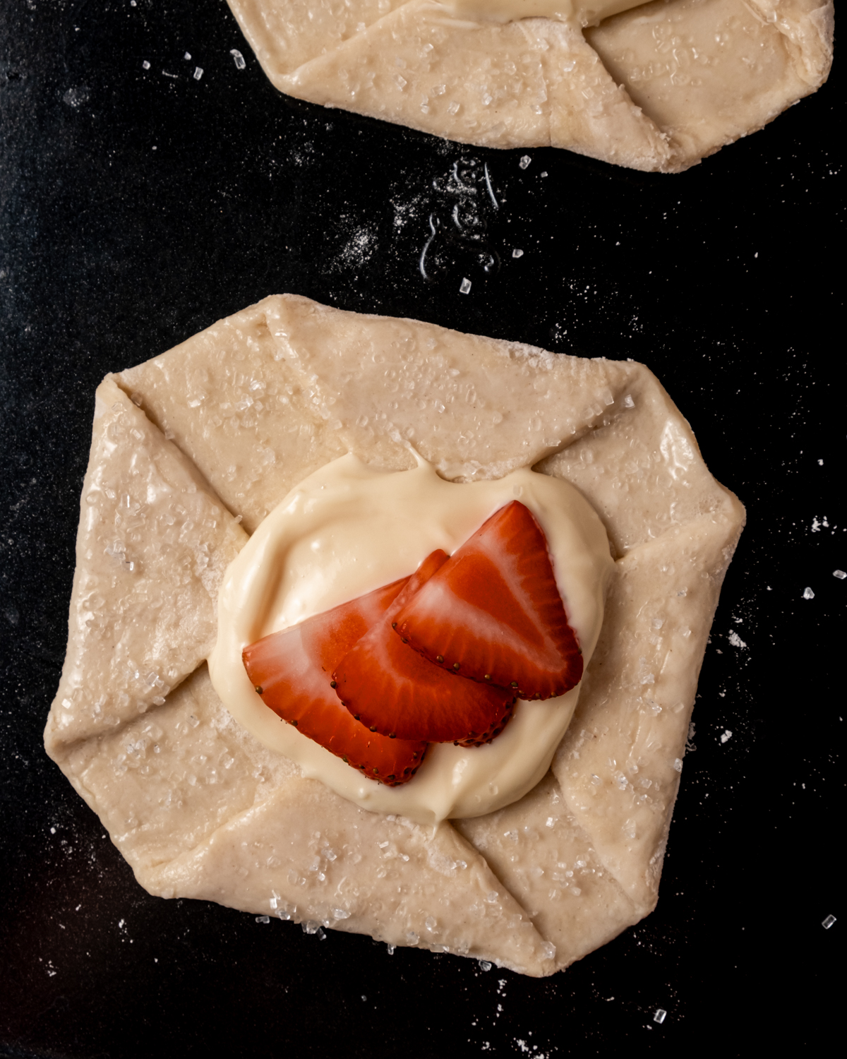 A close-up of an unbaked danish pastry with golden sugar-dusted puff pastry, filled with cream cheese mixture and topped with fresh sliced strawberries.