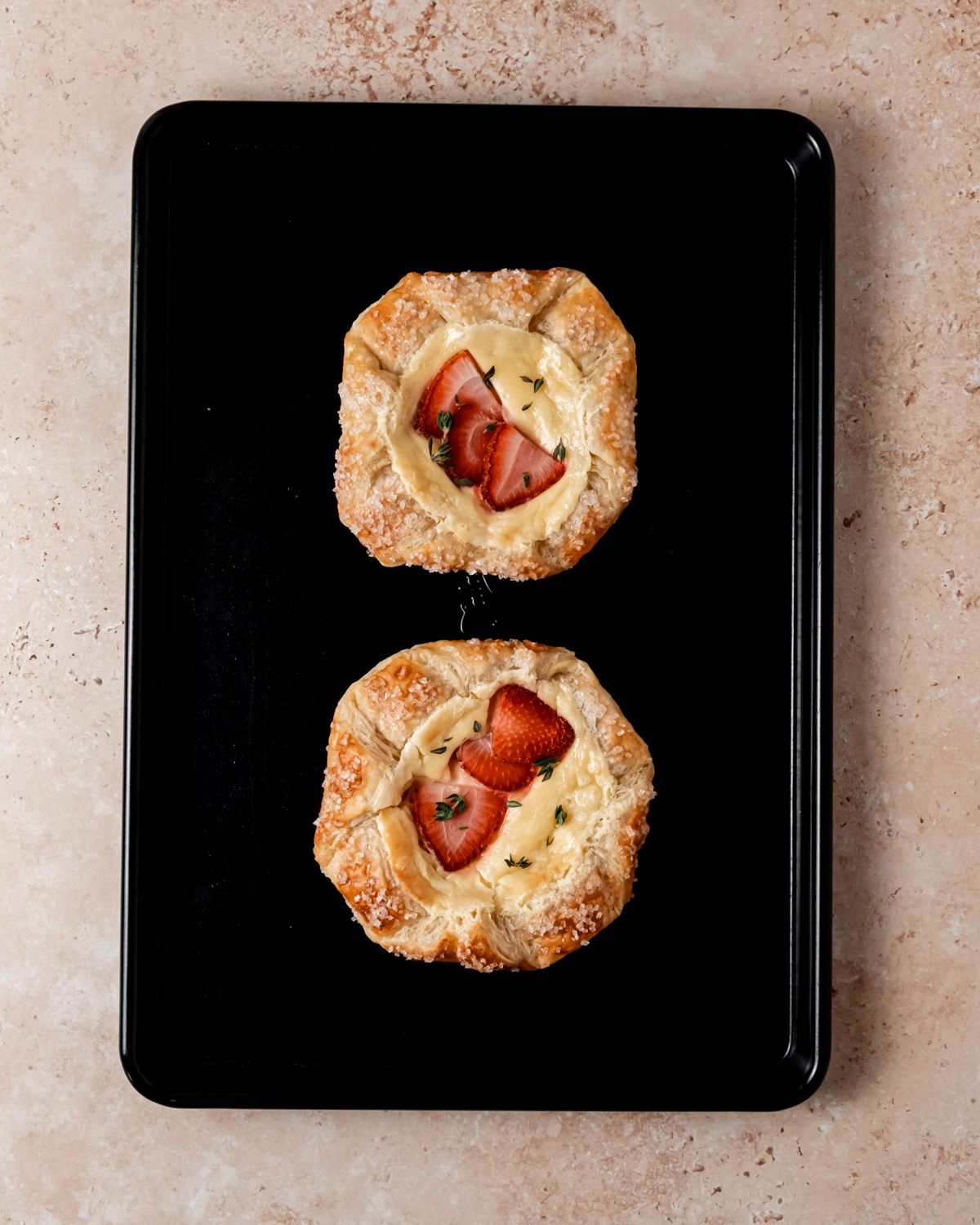 A top-down view of two baked strawberry cream cheese danishes on a black baking sheet, their golden brown pastry dusted with coarse sugar and garnished with fresh thyme.