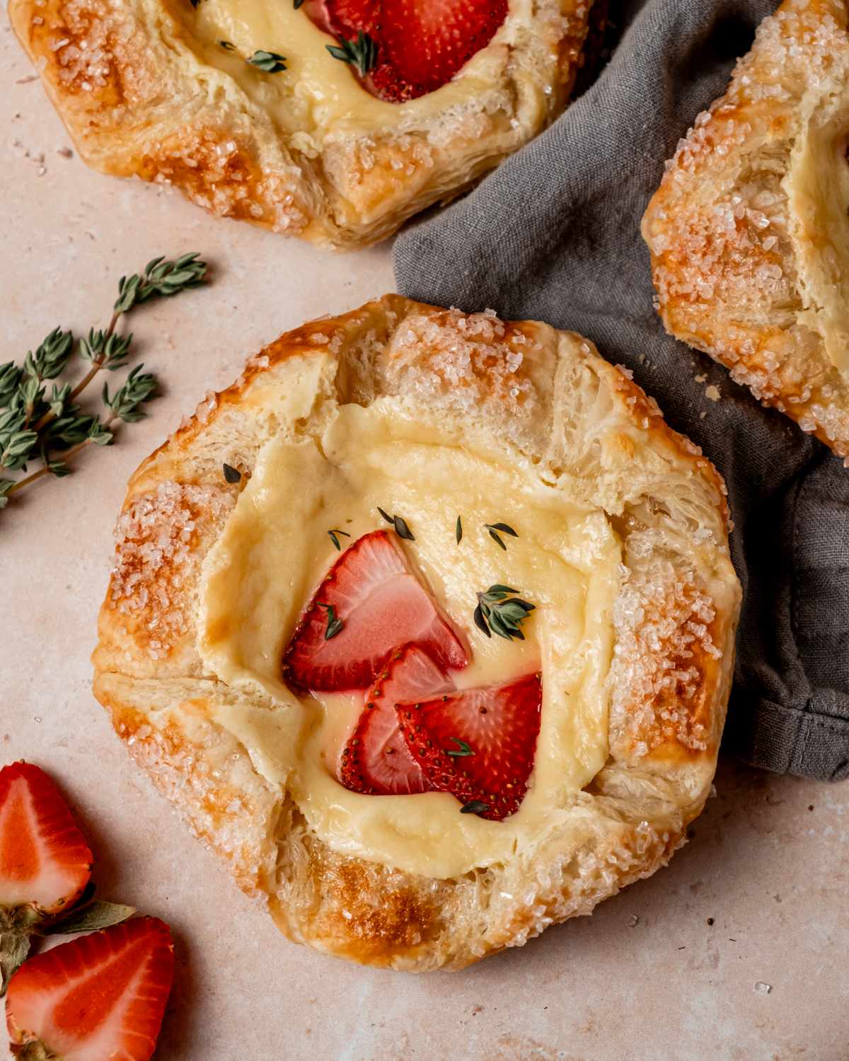 Close-up of a flaky strawberry Danish pastry topped with sliced strawberries and a few thyme leaves on a light beige surface, partially resting on a gray linen napkin.