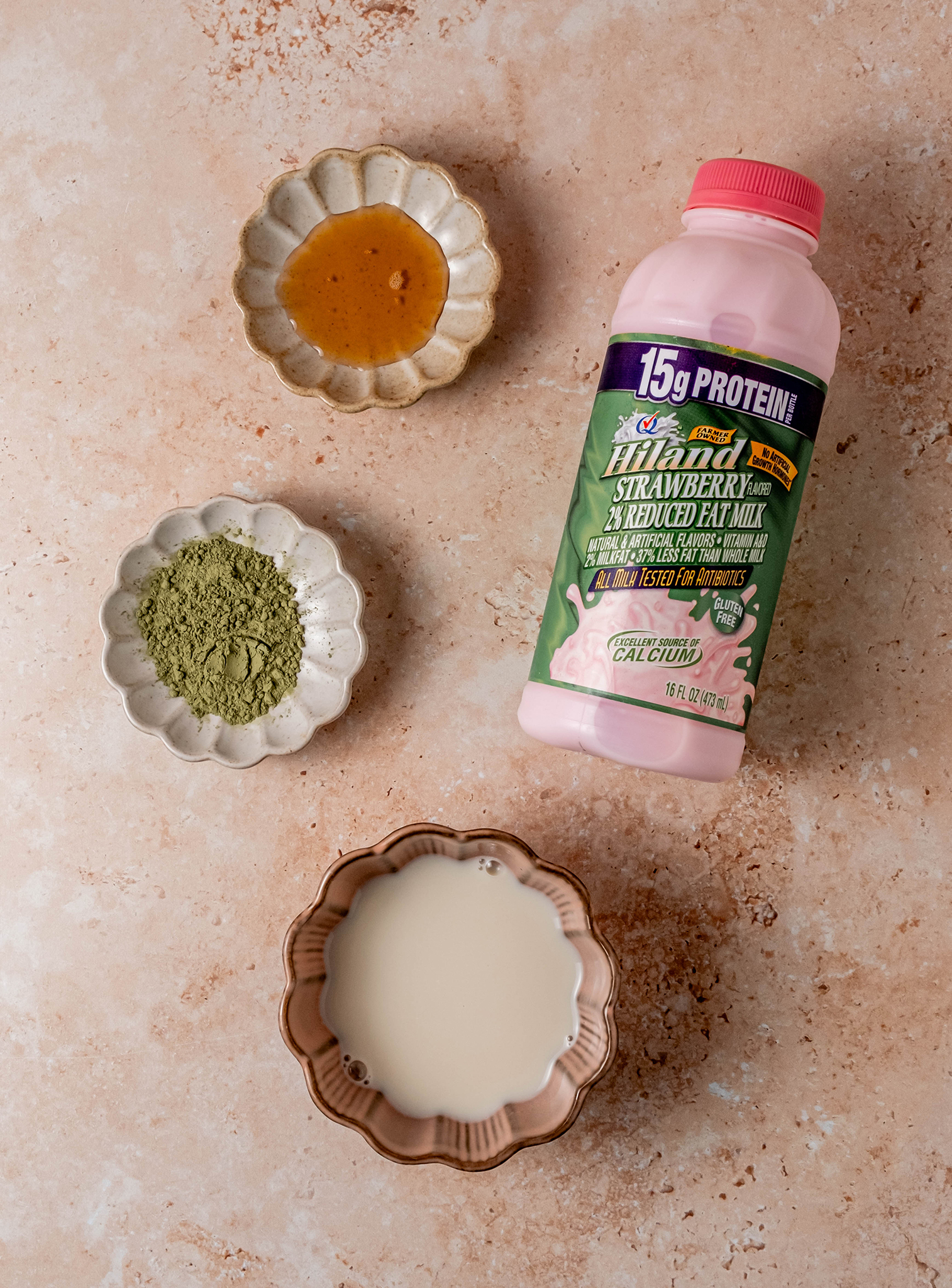 A flat lay of ingredients for a strawberry matcha latte, including a bowl of honey, a bowl of matcha powder, a bowl of milk, and a bottle of Hiland Strawberry Milk, all arranged on a peach-toned surface.