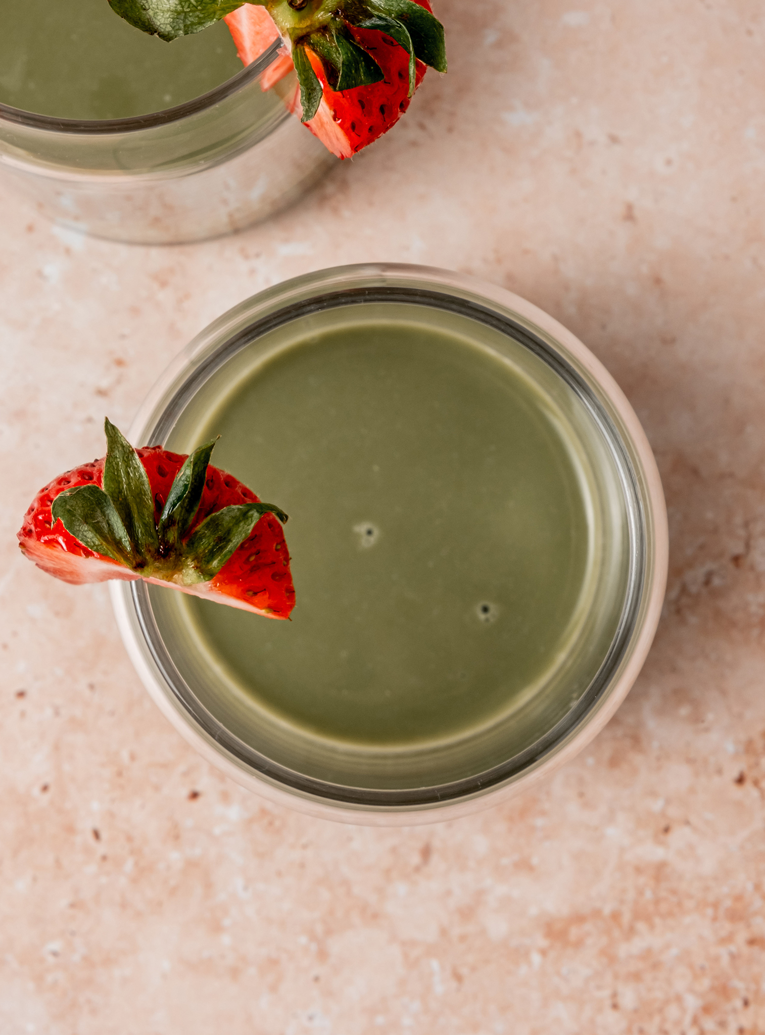 A glass of green matcha latte garnished with a sliced fresh strawberry on the rim, viewed from above on a neutral marble surface.