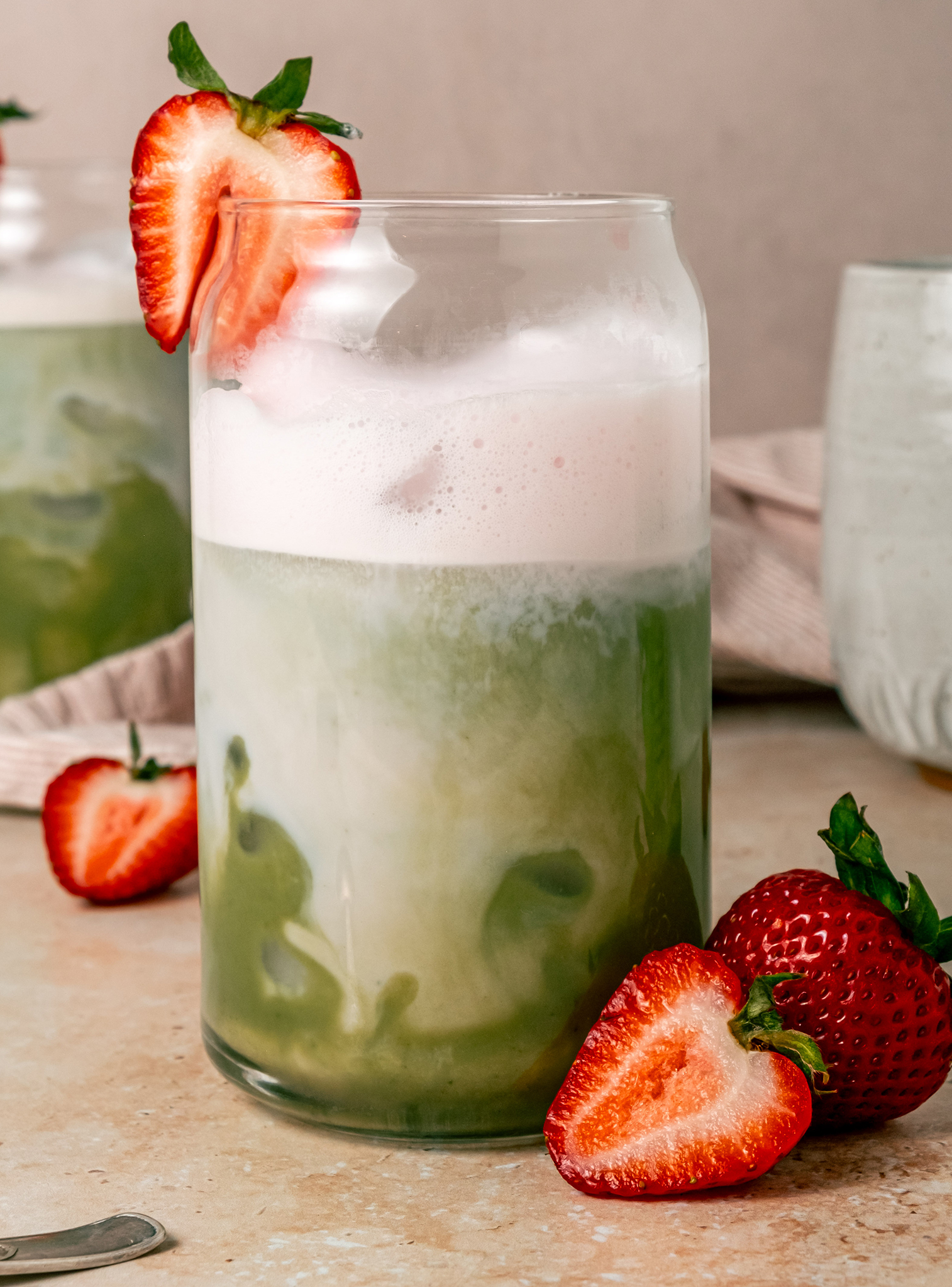 A close-up of a strawberry matcha latte in a clear glass, with green matcha and white foam layers swirling together, garnished with a halved strawberry and surrounded by more fresh strawberries.