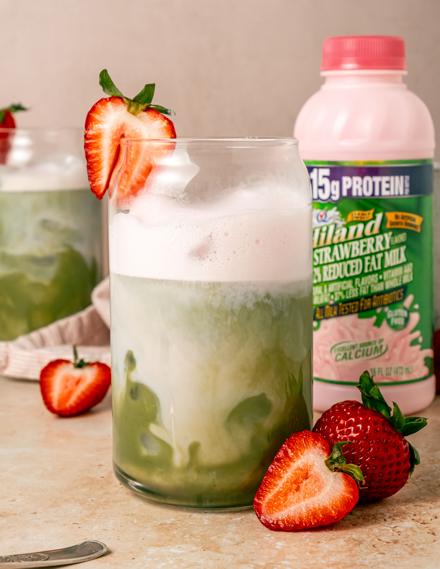 A close-up of a glass of iced strawberry matcha latte topped with pink foam and garnished with a fresh strawberry on the rim, with a bottle of Hiland Strawberry Milk and more strawberries in the background.