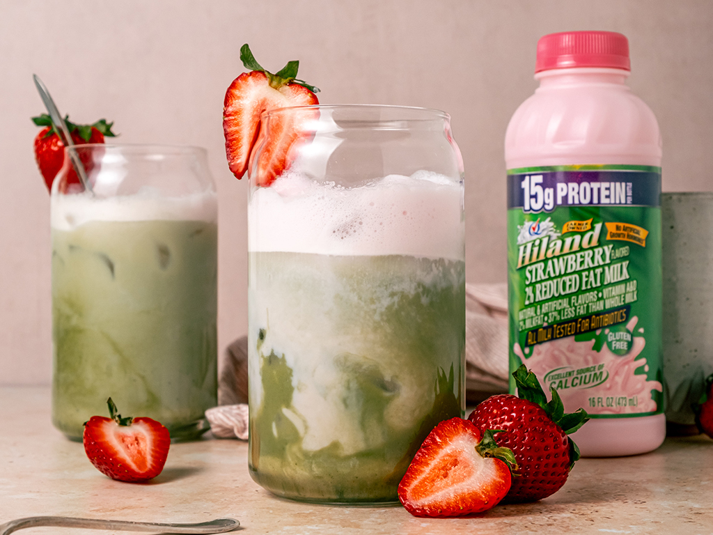 Two glasses of iced strawberry matcha latte with swirls of green matcha and pink milk, topped with frothy foam and garnished with halved strawberries, with a bottle of Hiland Strawberry Milk placed behind them.