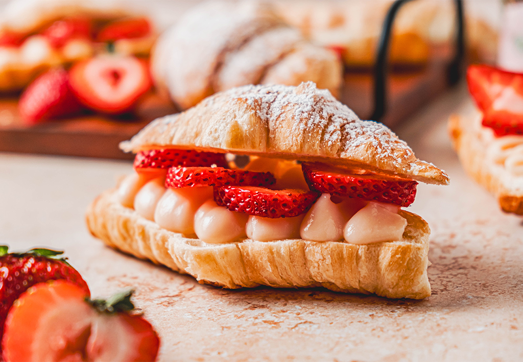 Flaky croissant filled with pale pink strawberry pastry cream and sliced fresh strawberries, lightly dusted with powdered sugar on a neutral surface.