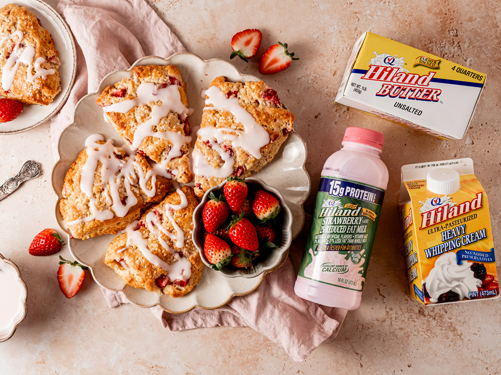 A plate of golden-brown strawberry scones drizzled with white icing, surrounded by fresh strawberries. A carton of Hiland Heavy Whipping Cream, a bottle of Hiland Strawberry Flavored 2% Reduced Fat Milk, and a box of Hiland Butter are arranged nearby on a beige surface with a soft pink cloth.