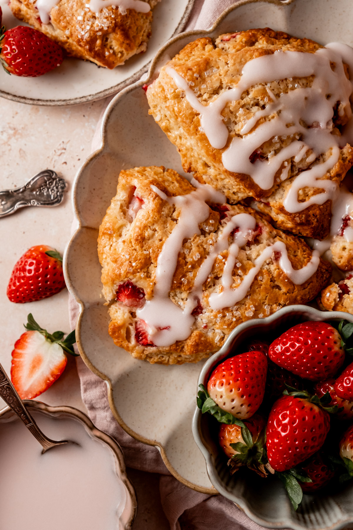 Overhead view of strawberry scones showcasing their flaky texture and juicy strawberry pieces. The scones are drizzled with a glossy white glaze, and a bowl of bright red fresh strawberries sits beside them.