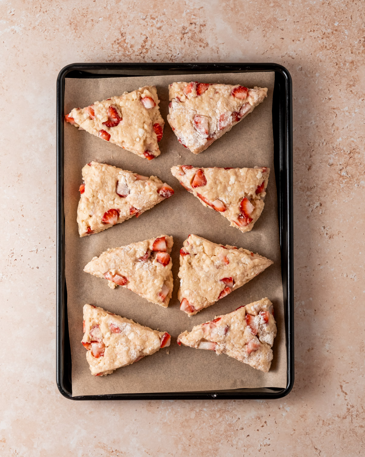 A black baking sheet lined with parchment paper, holding freshly cut, unbaked strawberry scone dough wedges.