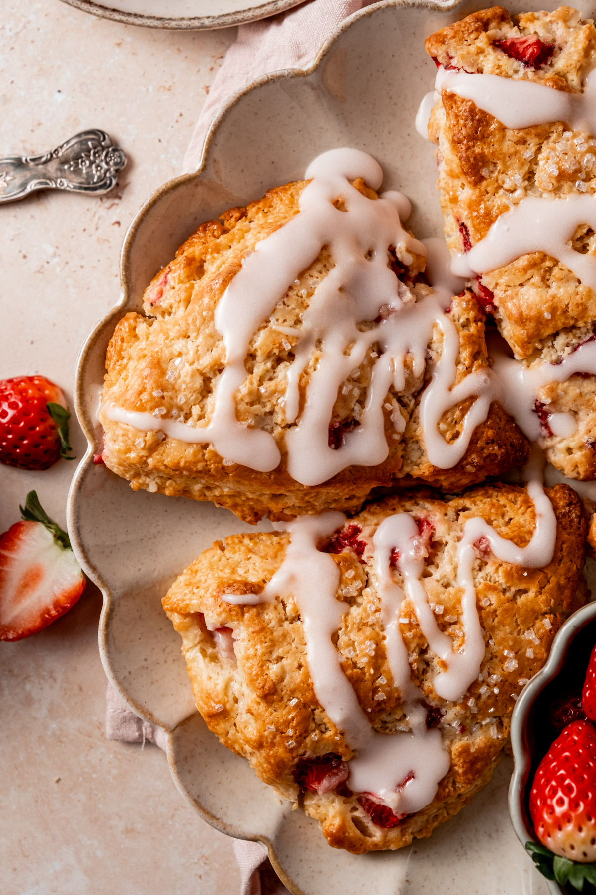 A close-up of golden strawberry scones drizzled with white glaze, arranged on a scalloped serving plate with fresh strawberries on the side.