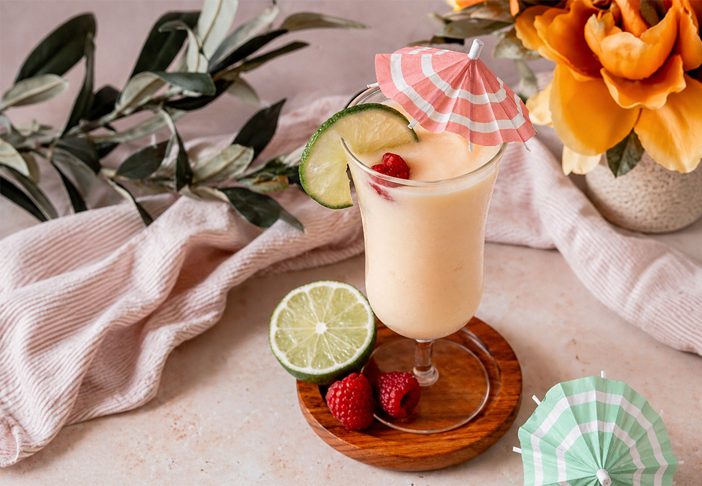 Tropical raspberry lemonade mocktail in a stemmed glass with a lime slice, raspberries, and a pink paper umbrella, set on a wooden coaster with fresh citrus and florals