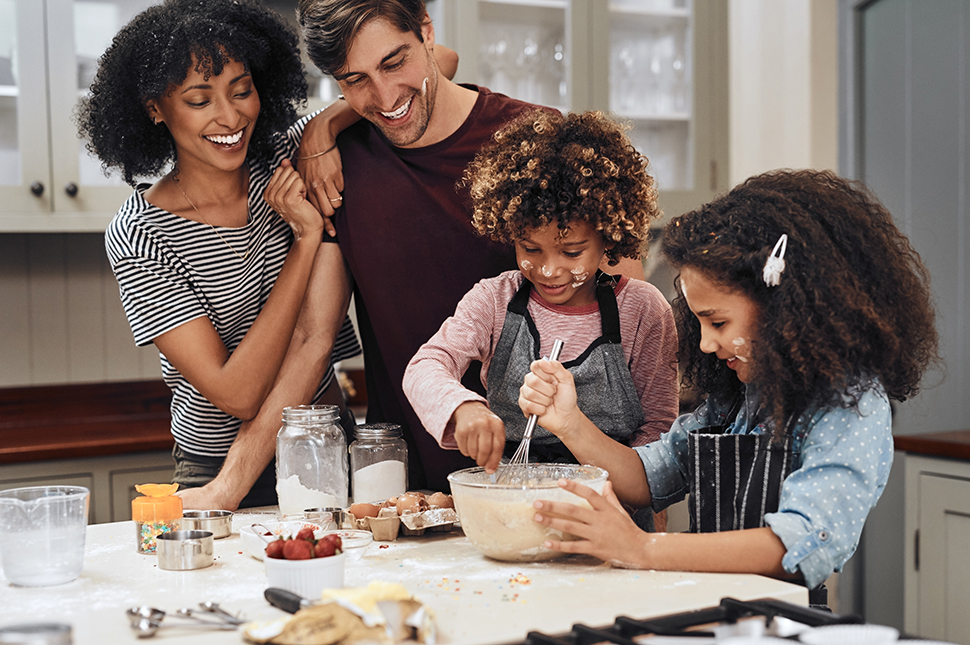 A joyful family of four bakes together in a cozy kitchen. The parents, smiling warmly, watch as their two young children mix ingredients in a large bowl. Baking supplies like flour, eggs, and measuring cups are spread across the counter, adding to the playful, hands-on atmosphere.