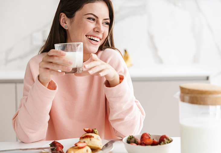 A young woman with long brown hair and a bright smile enjoys a glass of milk at a white kitchen table. She wears a soft pink sweatshirt and appears happy as she looks off to the side. In front of her is a plate of pastries topped with strawberries and a bowl of fresh strawberries, with a glass pitcher of milk nearby.